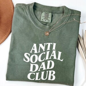Puede incluir: Una camiseta verde oliva con el texto "ANTI SOCIAL DAD CLUB" en blanco. Un collar dorado con un colgante circular descansa sobre la camiseta. La camiseta tiene una etiqueta Comfort Colors.