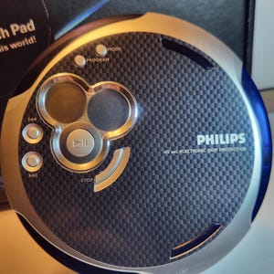 May include: A silver and blue Philips portable CD player with a carbon fiber-like texture. It features control buttons for mode, program, and playback, with the text "PHILIPS" and "45 sec ELECTRONIC SKIP PROTECTION" visible.
