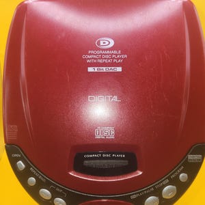 May include: A burgundy compact disc player with a black trim. The player has a digital display and several control buttons, including "Open," "Repeat/Rnd," and "Skip." The text "Programmable Compact Disc Player with Repeat Play" is visible on the device.