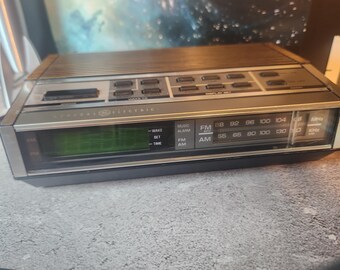 May include: A rectangular General Electric digital alarm clock radio with a wood-grain top panel and a silver-toned front face. It features a green digital display, a manual analog radio tuning dial for AM and FM frequencies, and multiple push buttons.