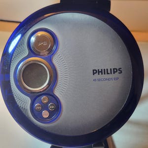 May include: A blue and silver Philips portable CD player. The device has a circular design with a play button, a display screen, and other control buttons. The text "PHILIPS" and "45 SECONDS ESP" are printed on the device.