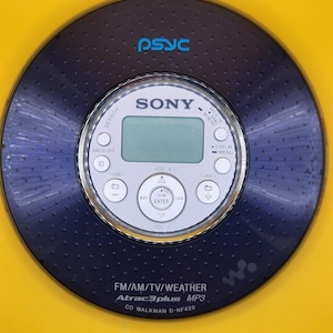 May include: A dark blue Sony CD Walkman D-NF420 player with a circular design. It features a central display, control buttons, and the text "SONY" and "PSYC". The player also has the text "FM/AM/TV/WEATHER Atrac3plus MP3".