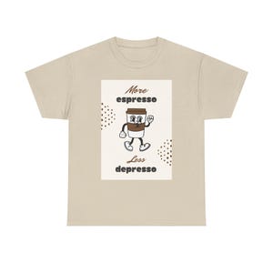 May include: A beige t-shirt with a rectangular graphic. The graphic shows a cartoon coffee cup character with the text "More espresso, Less depresso". Small brown dots are on either side of the graphic.