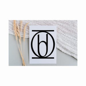 Può includere: Una carta bianca con un design monogramma nero stilizzato. Il design include un cerchio, linee orizzontali e una forma di lettera centrale. La carta è posta su uno sfondo neutro con spighe di grano secche e tessuto bianco.