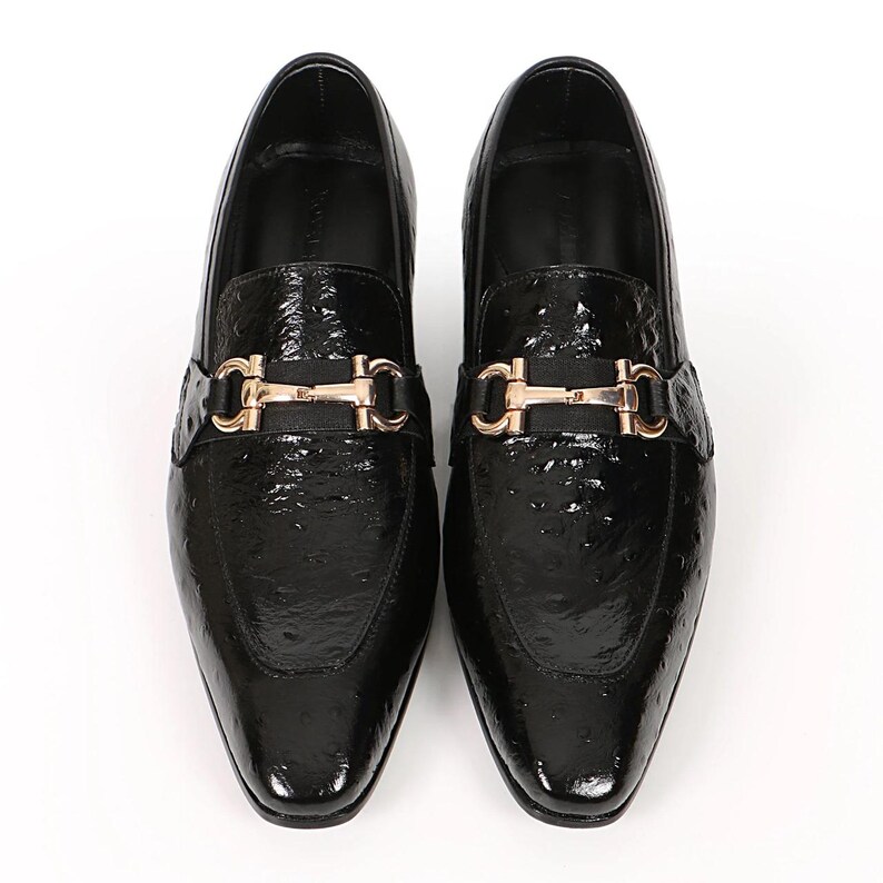 May include: Pair of black leather loafers with a textured finish. Each shoe features a gold-colored buckle detail across the vamp. The shoes have a square toe and a glossy appearance, suitable for formal wear.