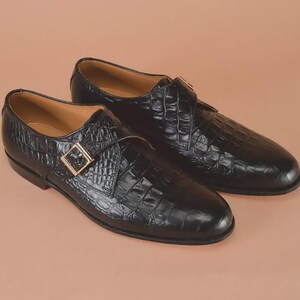 May include: A pair of black leather dress shoes with a crocodile texture. The shoes feature a buckle closure with a gold-colored buckle. The soles are black with a red accent. The shoes are displayed on a neutral background.