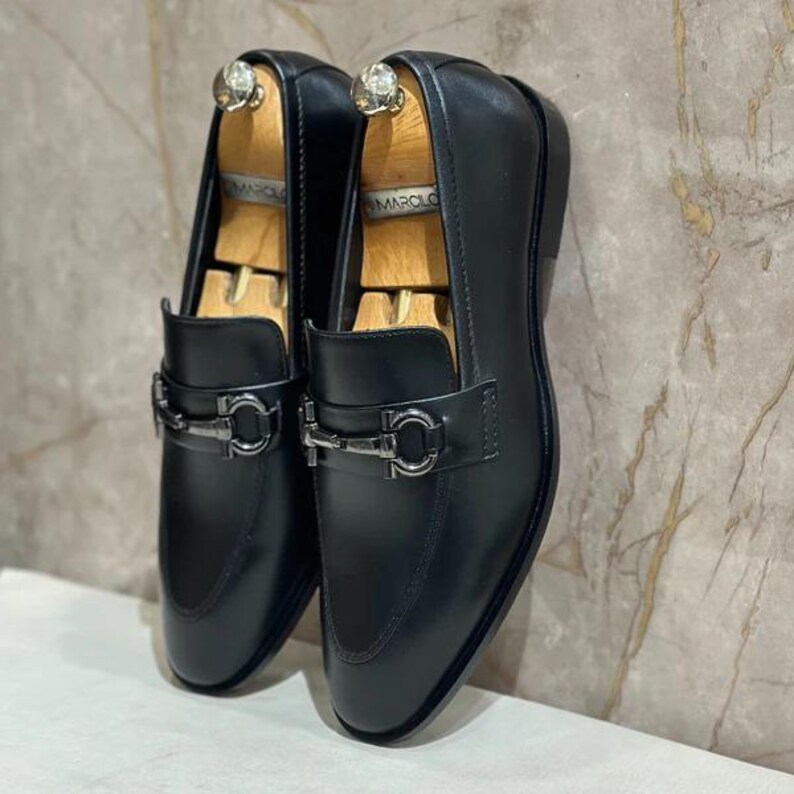 May include: A pair of black leather loafers with silver-tone metal buckles. The shoes are displayed with wooden shoe trees inside. The loafers have a classic design, suitable for formal or semi-formal occasions.