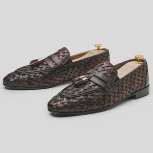 May include: A pair of brown woven leather loafers with tassel accents. The shoes have a classic design with a woven pattern and a leather strap across the top. The loafers are displayed on shoe trees.