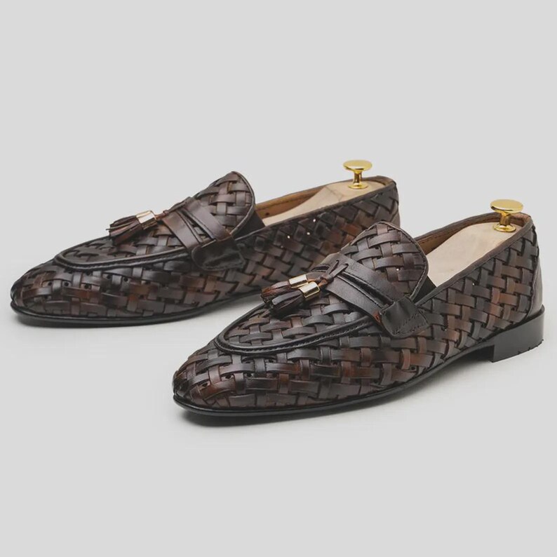 May include: A pair of brown woven leather loafers with tassel accents. The shoes have a classic design with a woven pattern and a leather strap across the top. The loafers are displayed on shoe trees.