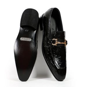 May include: Pair of black leather loafers with a glossy finish. The shoes feature a gold-colored buckle detail on the vamp. The soles are black, and one shoe is turned to show the sole and the "100% Leather Handmade" label.