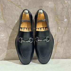 May include: A pair of black leather loafers with silver-tone hardware. The shoes have a classic slip-on design with a metal buckle detail across the vamp. Wooden shoe trees are inserted inside each shoe.