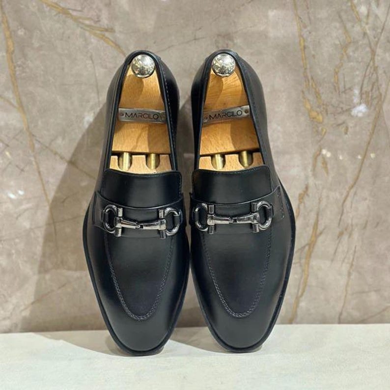 May include: A pair of black leather loafers with silver-tone hardware. The shoes have a classic slip-on design with a metal buckle detail across the vamp. Wooden shoe trees are inserted inside each shoe.