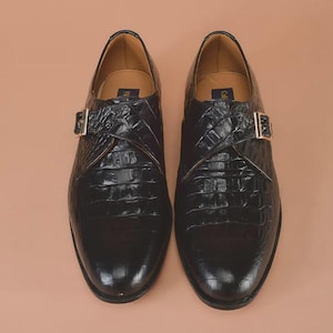May include: A pair of black leather dress shoes with a crocodile skin texture. Each shoe has a gold-colored buckle closure on the side. The shoes are displayed on a light brown surface.