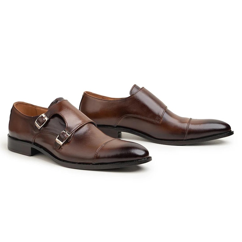 May include: A pair of brown leather dress shoes with a double monk strap closure. The shoes have a polished finish and a pointed toe. The buckles are silver-colored. The shoes are displayed on a white background.