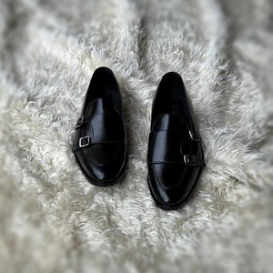 May include: A pair of black leather monk strap shoes with silver buckles. The shoes are displayed on a white, fluffy surface. The shoes have a glossy finish and a classic design.