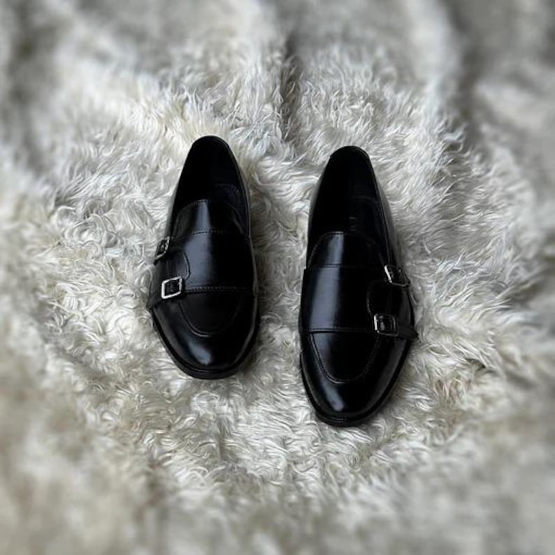 May include: A pair of black leather monk strap shoes with silver buckles. The shoes are displayed on a white, fluffy surface. The shoes have a glossy finish and a classic design.