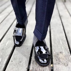May include: Black patent leather double monk strap shoes with silver buckles. The shoes have a glossy finish and are paired with dark blue trousers and gray socks. The image is taken from a low angle, showing the shoes on a wooden surface.