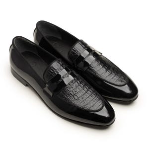 May include: A pair of glossy black leather loafers. The shoes have a patent finish and a textured, crocodile-like pattern on the vamp. The design includes a strap across the top. The shoes are displayed on a white background.
