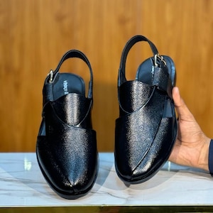 Op de afbeelding: Paar zwarte leren sandalen met een slingback-band en een getextureerde afwerking. De sandalen hebben een gesloten teenontwerp en een band over de bovenkant. Het woord "Vibes" is zichtbaar op de binnenzool. De sandalen worden gepresenteerd op een marmeren oppervlak.