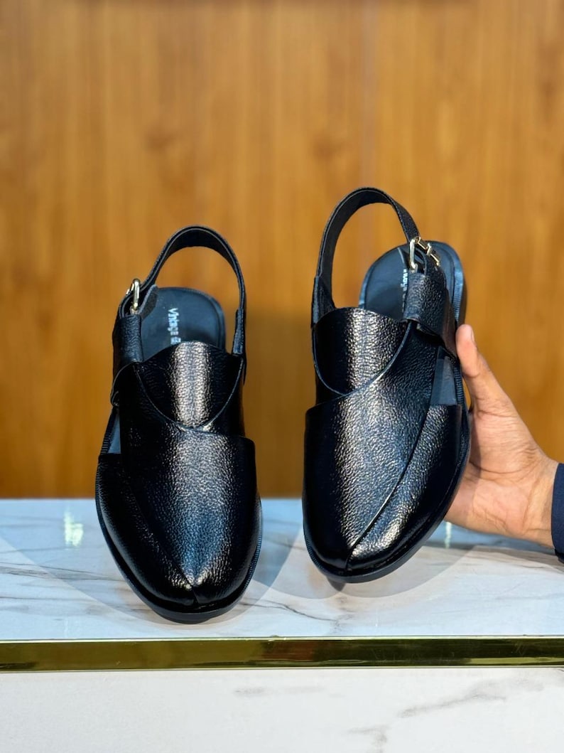 Op de afbeelding: Paar zwarte leren sandalen met een slingback-band en een getextureerde afwerking. De sandalen hebben een gesloten teenontwerp en een band over de bovenkant. Het woord "Vibes" is zichtbaar op de binnenzool. De sandalen worden gepresenteerd op een marmeren oppervlak.
