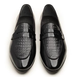 May include: A pair of glossy black loafers. The shoes have a patent leather finish with a textured, crocodile-like pattern on the vamp. The design includes a strap across the top. The shoes are displayed on a white background.