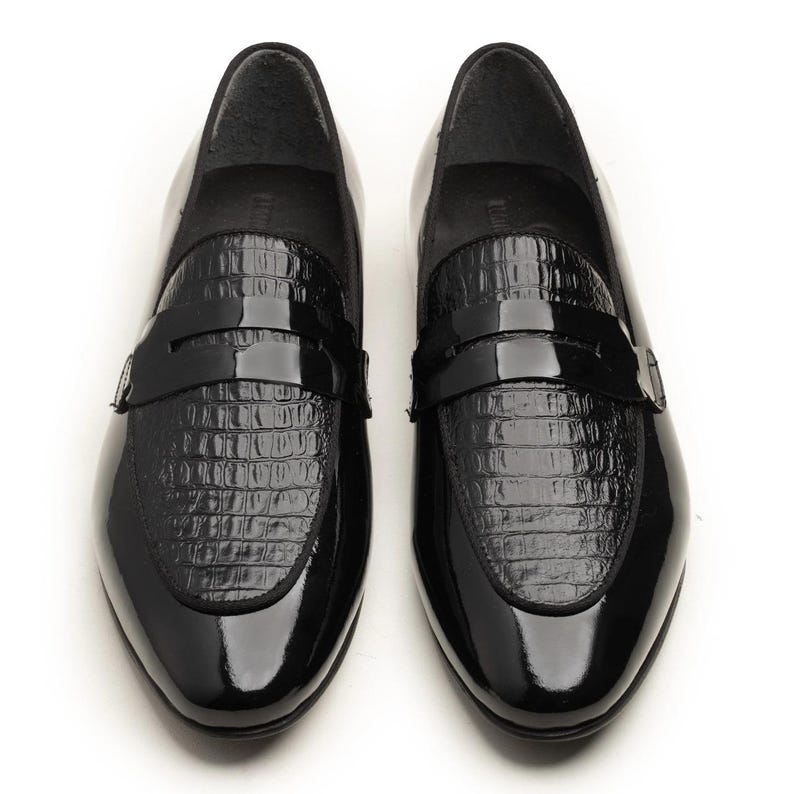 May include: A pair of glossy black loafers. The shoes have a patent leather finish with a textured, crocodile-like pattern on the vamp. The design includes a strap across the top. The shoes are displayed on a white background.