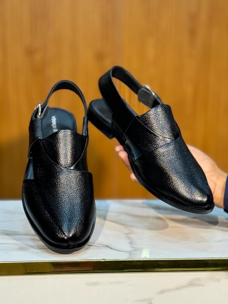 Op de afbeelding: Twee zwarte leren sandalen met een getextureerde afwerking. De sandalen hebben een slingback-band met een gesp en een gesloten teenontwerp. De schoenen worden gepresenteerd op een marmeren oppervlak.