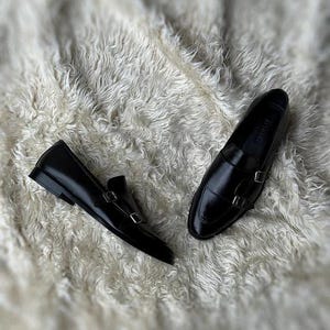 May include: A pair of black leather double monk strap shoes. The shoes have a shiny finish and silver buckles. They are displayed on a white, textured surface.