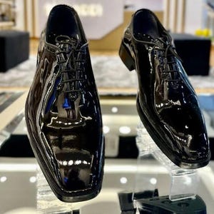May include: Two black patent leather dress shoes with a high-gloss finish. The shoes have a classic lace-up design and a square toe. The image shows the shoes from a slightly elevated angle, highlighting their polished appearance and sleek silhouette.