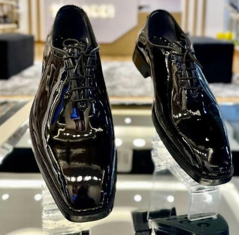 May include: Two black patent leather dress shoes with a high-gloss finish. The shoes have a classic lace-up design and a square toe. The image shows the shoes from a slightly elevated angle, highlighting their polished appearance and sleek silhouette.