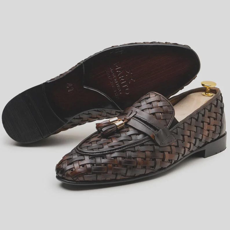 May include: Brown leather woven loafers with a tassel detail and a leather sole. The shoes are displayed with one shoe upright and the other showing the sole. The shoes have a classic design.