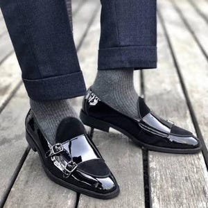 May include: Black patent leather double monk strap shoes with a suede vamp. The shoes are worn with gray socks and dark blue denim jeans. The shoes are on a wooden surface.