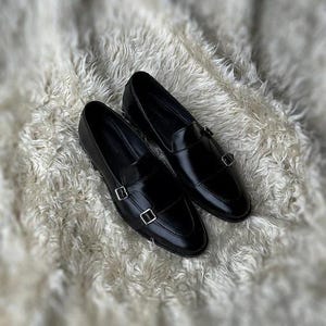 May include: A pair of shiny black leather loafers with silver-colored double monk straps. The shoes are displayed on a soft, white, textured surface. The loafers have a classic design, suitable for formal or semi-formal occasions.