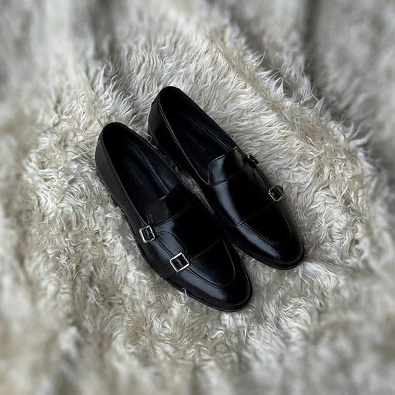 May include: A pair of shiny black leather loafers with silver-colored double monk straps. The shoes are displayed on a soft, white, textured surface. The loafers have a classic design, suitable for formal or semi-formal occasions.