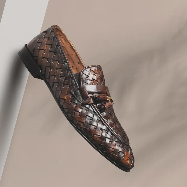 May include: A single brown leather loafer with a woven design. The shoe features a tassel detail and a dark sole. The leather has a rich, textured appearance, and the shoe is angled slightly, showcasing its design.