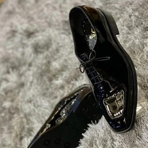 May include: Pair of glossy black leather dress shoes with a square toe and lace-up closure. The shoes have a patent leather finish and a decorative design on the toe. The soles are black and the shoes are set against a gray, textured background.