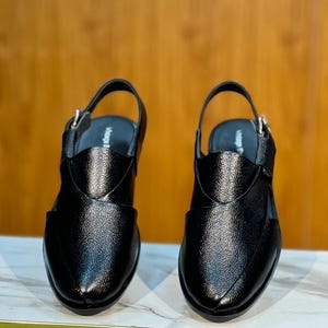Op de afbeelding: Een paar zwarte leren sandalen met een getextureerde afwerking. De sandalen hebben een slingback-band en een gespsluiting. Het ontwerp heeft een spitse neus en een decoratieve overlay op de wreef. De schoenen worden op een marmeren oppervlak weergegeven.
