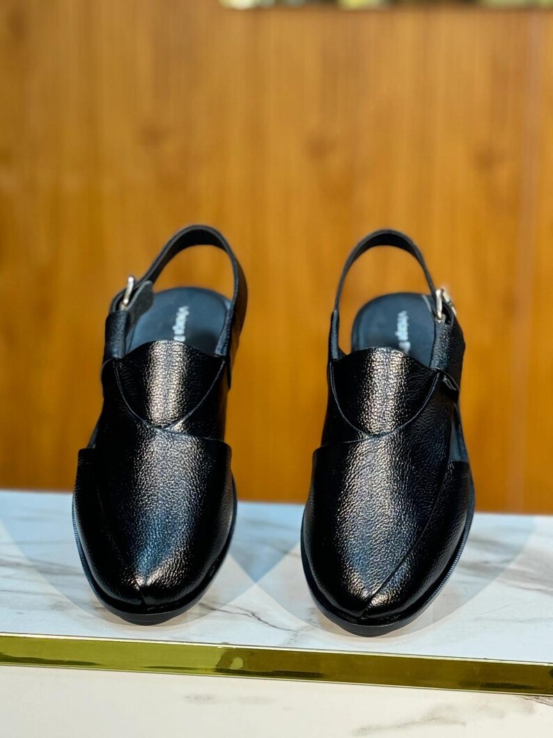 Op de afbeelding: Een paar zwarte leren sandalen met een getextureerde afwerking. De sandalen hebben een slingback-band en een gespsluiting. Het ontwerp heeft een spitse neus en een decoratieve overlay op de wreef. De schoenen worden op een marmeren oppervlak weergegeven.
