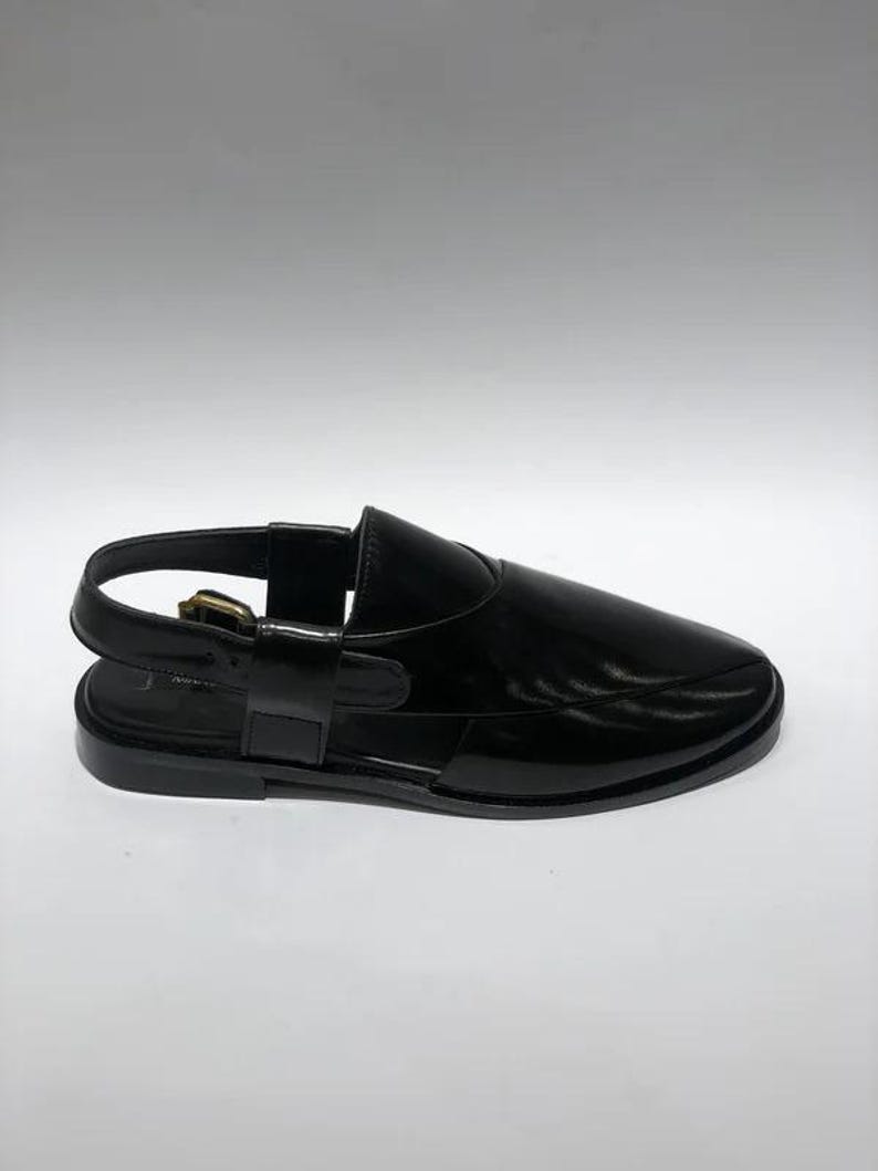 May include: Black leather sandal with a slingback strap and buckle closure. The sandal features a rounded toe and a flat sole. The shoe has a glossy finish and a minimalist design.