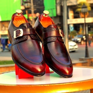 May include: A pair of burgundy leather dress shoes with buckle fastenings. The shoes have a polished finish and are displayed on a round table with a gold trim. The background is a blurred outdoor scene.