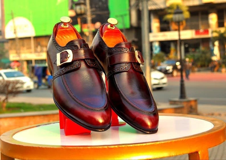 May include: A pair of burgundy leather dress shoes with buckle fastenings. The shoes have a polished finish and are displayed on a round table with a gold trim. The background is a blurred outdoor scene.