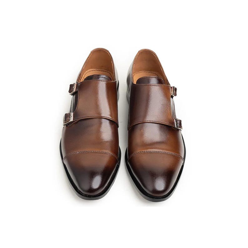 May include: A pair of brown leather dress shoes with double monk straps and silver buckles. The shoes have a polished finish and a classic design, perfect for formal occasions.