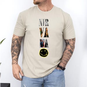 May include: Beige t-shirt featuring the band name "NIRVANA" in a vertical design with band member images within the letters. Below the text is a yellow smiley face with black eyes and a black mouth. The shirt is a neutral color.