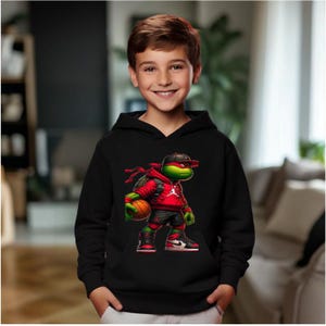 May include: A black hooded sweatshirt featuring a cartoon image of a Teenage Mutant Ninja Turtle wearing a red hoodie, black shorts, and sneakers, holding a basketball. The design includes the Jumpman logo. The child is smiling.