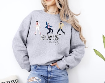 May include: A light gray sweatshirt featuring three cartoon illustrations of Elvis Presley in different poses. The word "ELVIS" is printed in a bold, black, and white font, with his signature below.