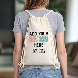 May include: A cream-colored drawstring backpack featuring the black text ADD YOUR DESIGN HERE, with the word DESIGN in a multi-colored gradient, and a rectangular box below containing the text TEXT - PHOTO IMAGE - LOGO in black.