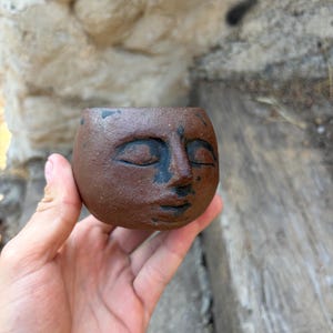 May include: A small, round, brown ceramic planter featuring a sculpted human face with closed eyes and a nose. The surface has a textured, matte finish with dark, charcoal-colored accents highlighting the facial features and contours.