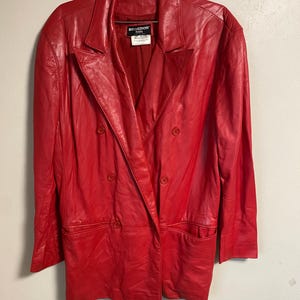May include: A vibrant red leather blazer with a double-breasted design. The jacket features a notched collar, two front pockets, and four buttons. The label reads "REGEDOR Italia". The blazer is hanging on a metal hanger.