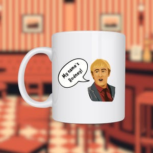 Può includere: Tazza in ceramica bianca con un'illustrazione a cartoni animati di un uomo con i capelli biondi e il testo "My name's Rodney!" in un fumetto. L'uomo indossa una giacca grigia e una camicia rossa.