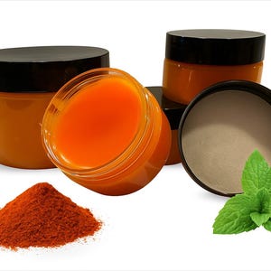 Op de afbeelding: Verschillende kleine, transparante potjes gevuld met een oranje substantie, elk met een zwarte deksel. Een hoop rood poeder en een takje groene munt zijn ook aanwezig. De potjes staan op een witte achtergrond.